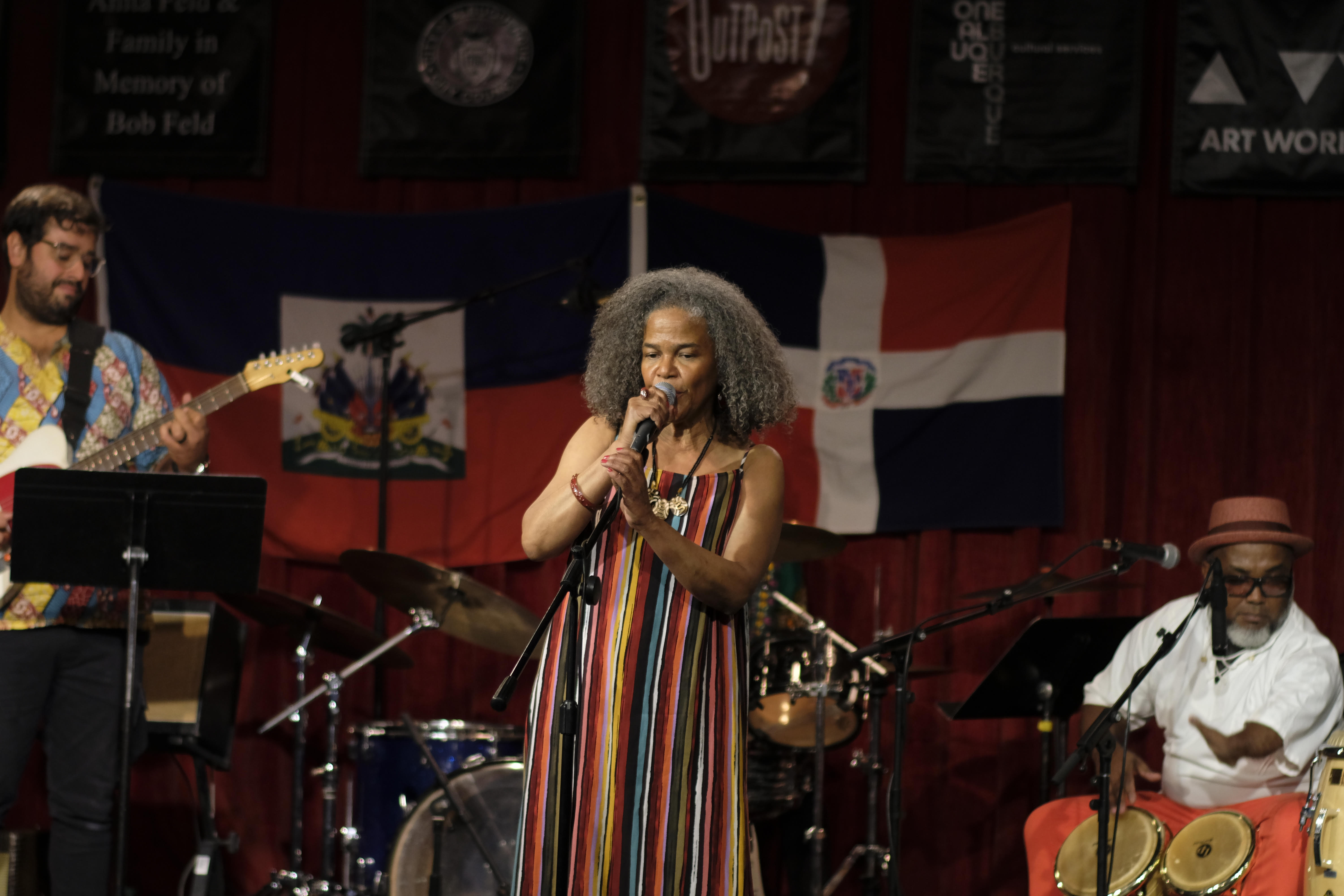 Maria Terrero of Kumba Carey in a multi-colored stripe dress gripping a microphone