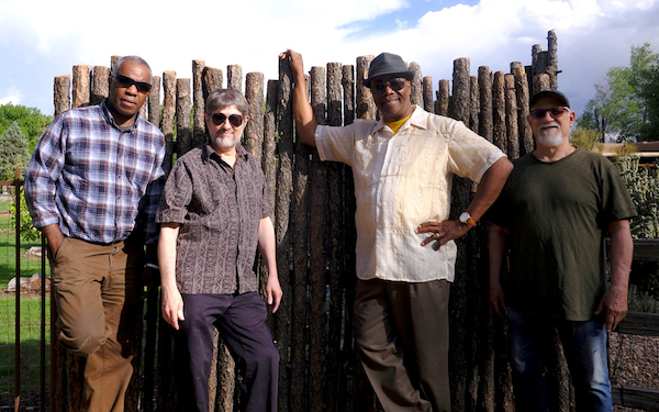 Four members of Racine Kreyol wearing sunglassed leaned up against a wooden fence Bright sky