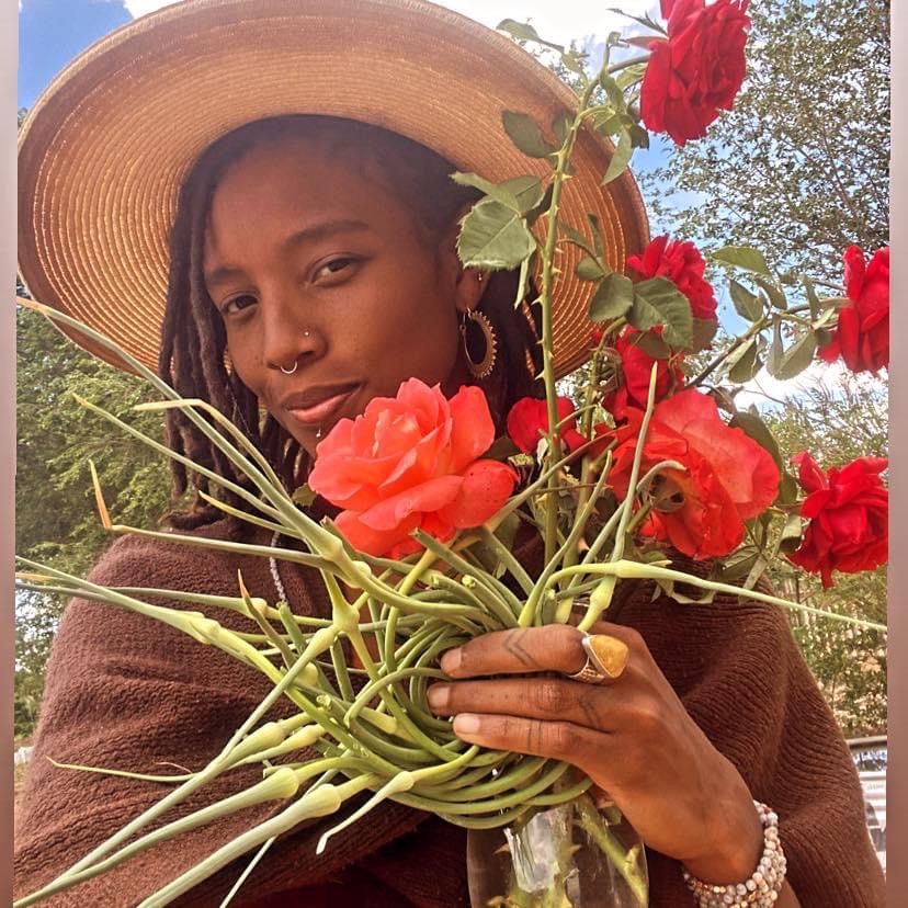 woman in a wide-brimmed straw hat and a nose ring holding garlic scapes and large bright coral roses