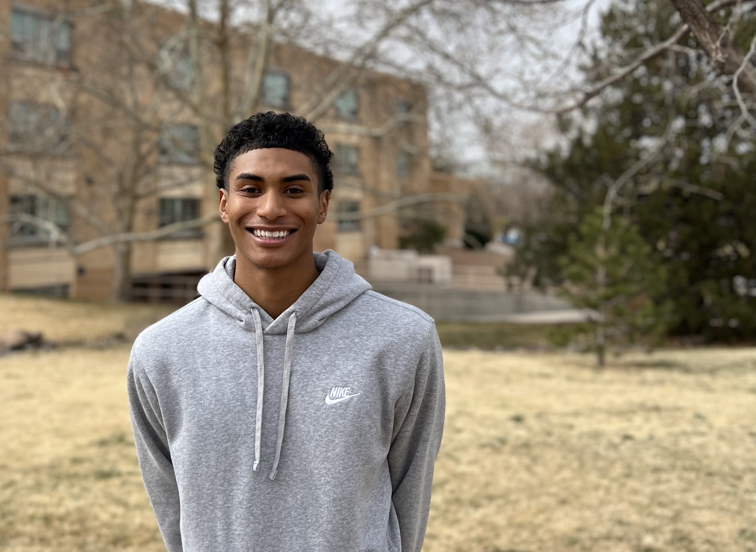 smiling young man in a grey hoodie against a backdrop of a brick building and pine trees