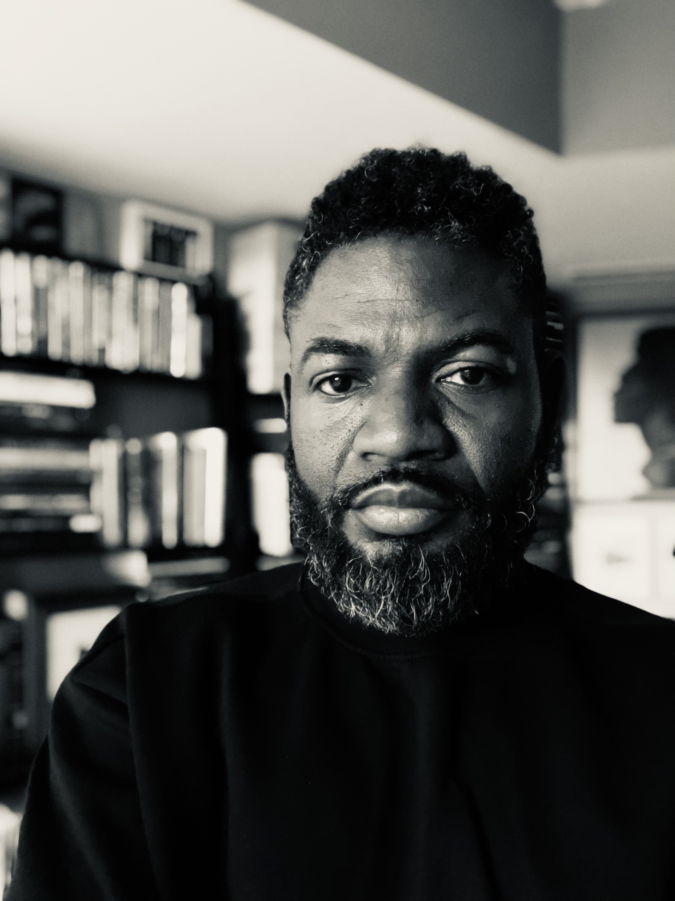 black and white photo of a bearded man with blurred background of what appear to be books